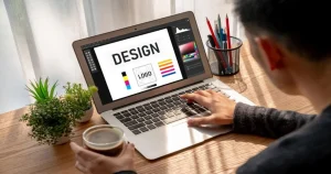 A person using a laptop with a design program open, holding a coffee cup, and checking social media